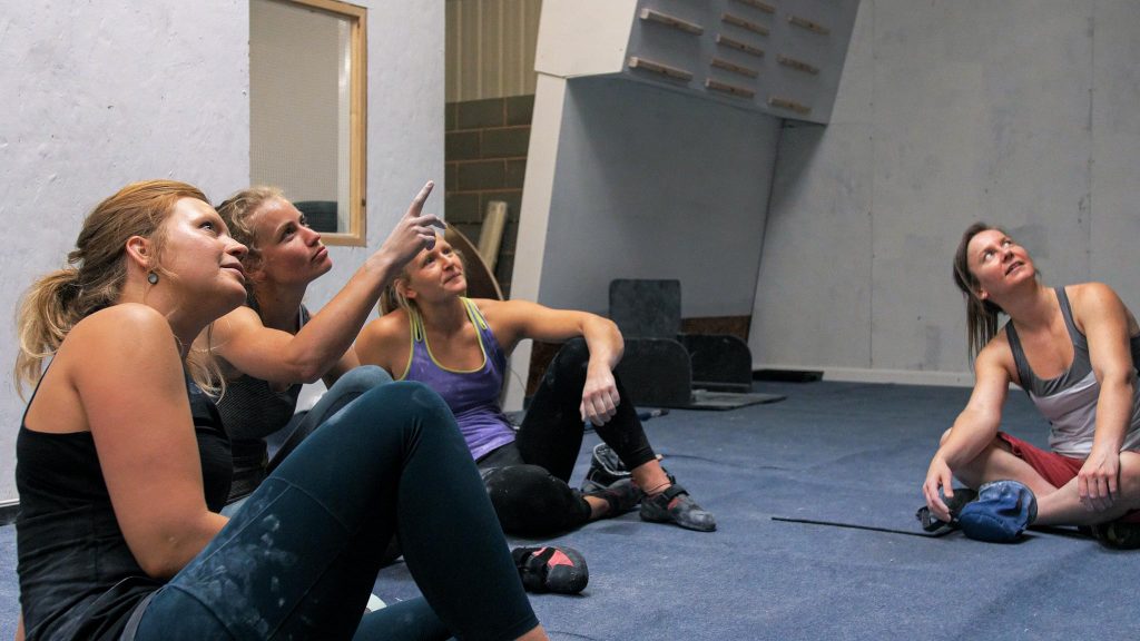 Four women sit on the mats in a bouldering gym, looking at the climbing holds on the wall. The woman in the middle points at the wall.